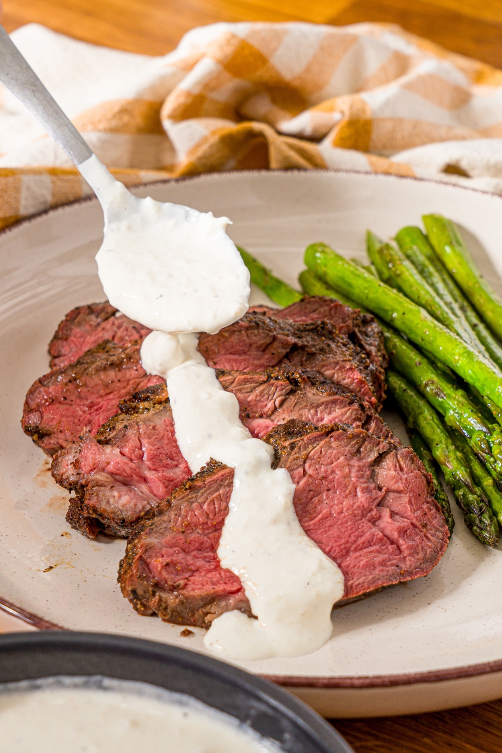 A white plate with sliced steak and asparagus, with a spoon pouring horseradish sauce over the steak. The plate is on a wooden counter with a yellow checkered napkin.
