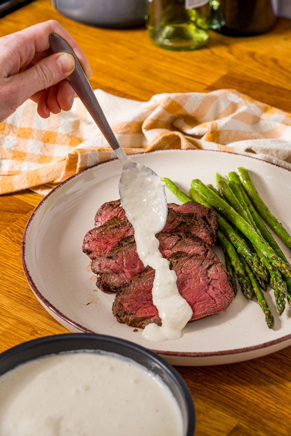 A white plate with sliced steak and asparagus, with a spoon pouring horseradish sauce over the steak. The plate is on a wooden counter with a yellow checkered napkin.