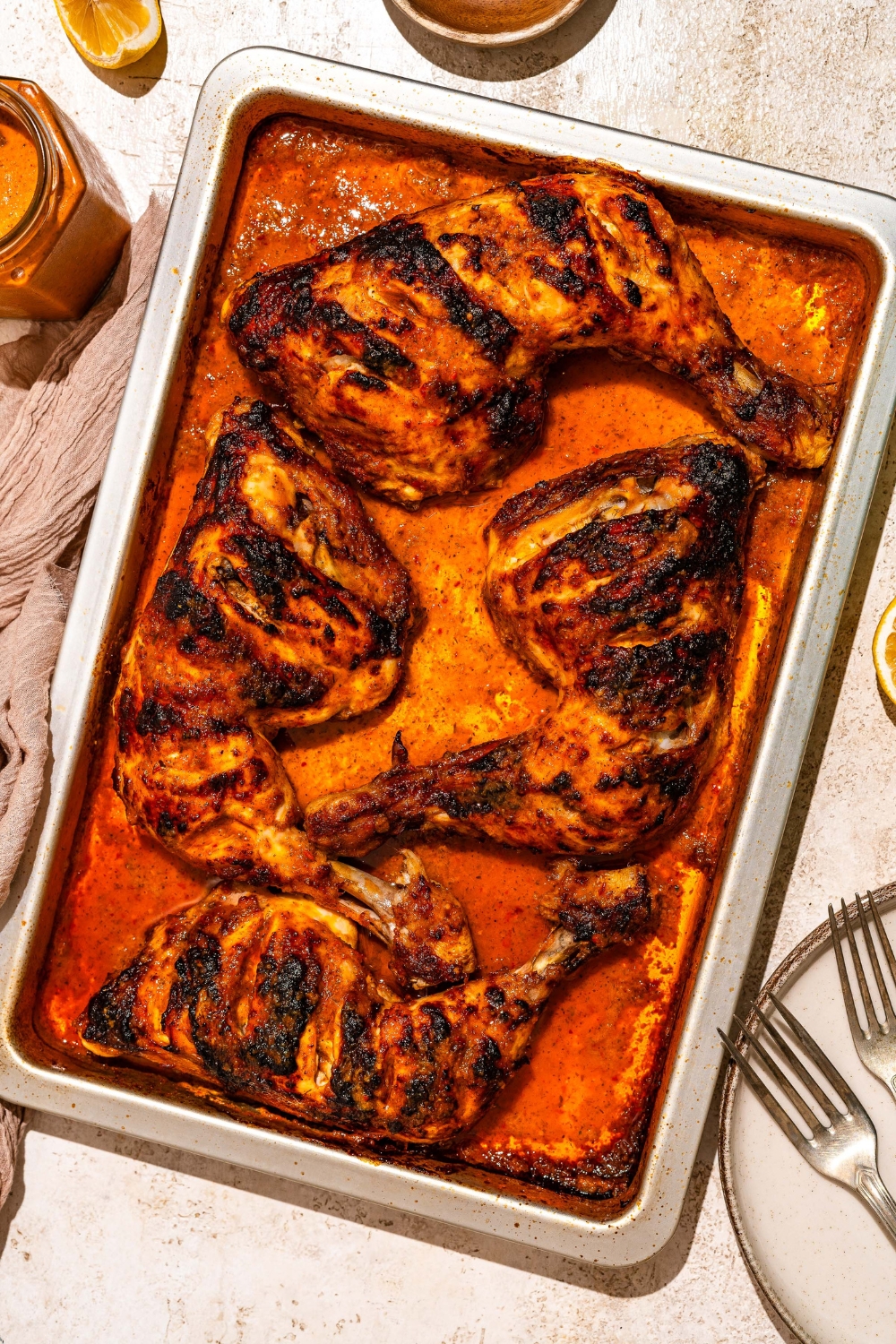 A sheet pan with roasted chicken cooked in peri peri sauce. The pan is on a tan counter with a jar of sauce.