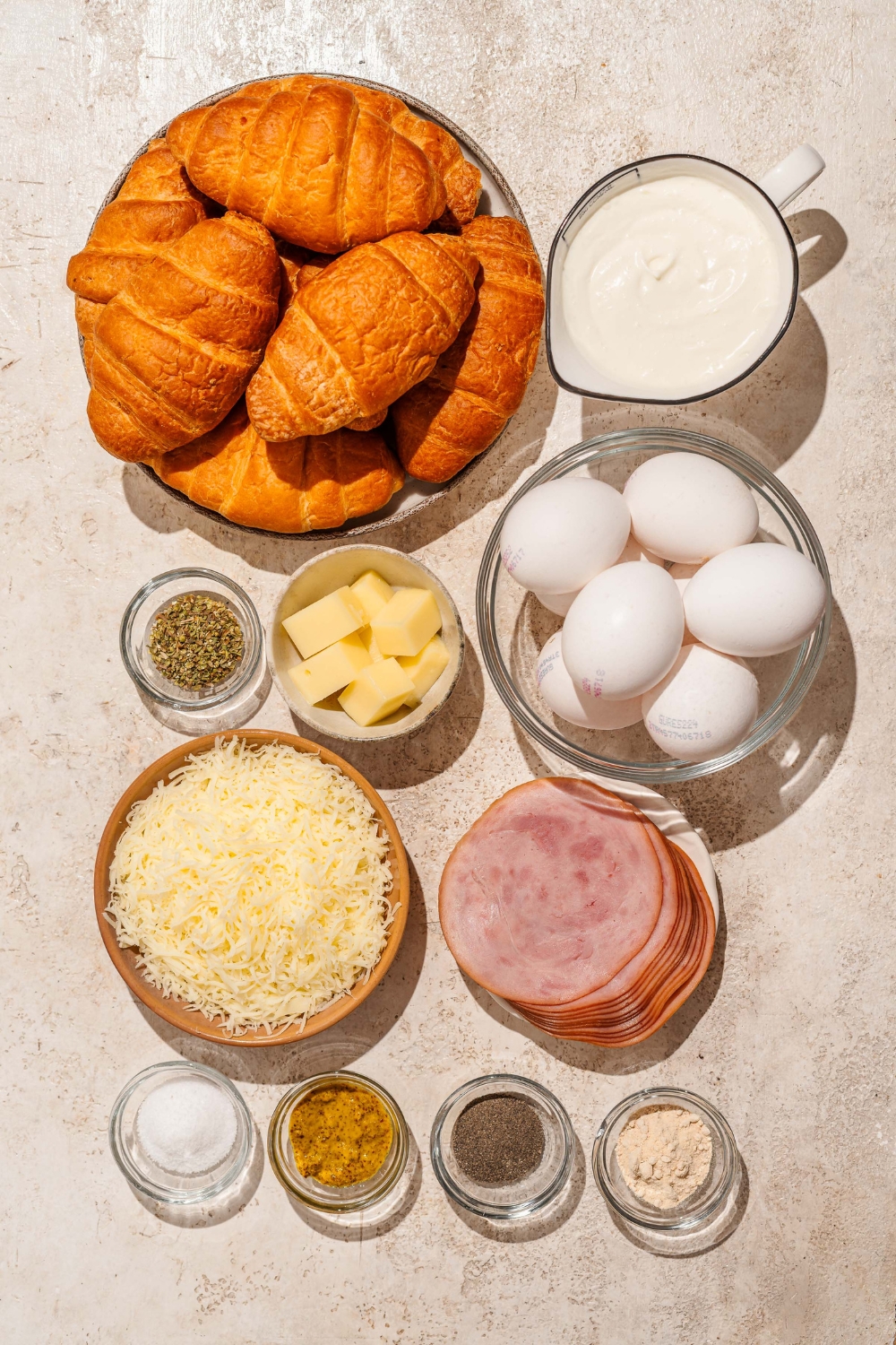 A tan counter with several bowls containing ingredients to make croissant breakfast casserole including croissants, eggs, ham, cheese, heavy cream, mustard, and seasonings.
