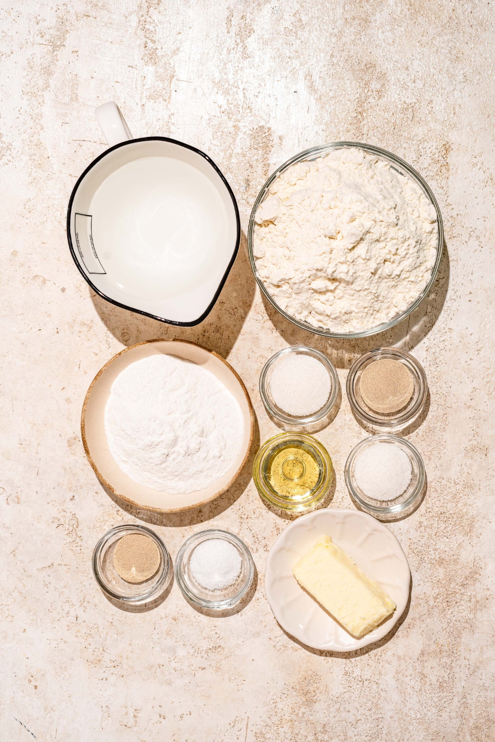 A tan counter with several bowls containing ingredients to make Dutch Crunch bread including instant yeast, water, butter, oil, flour, and sugar.