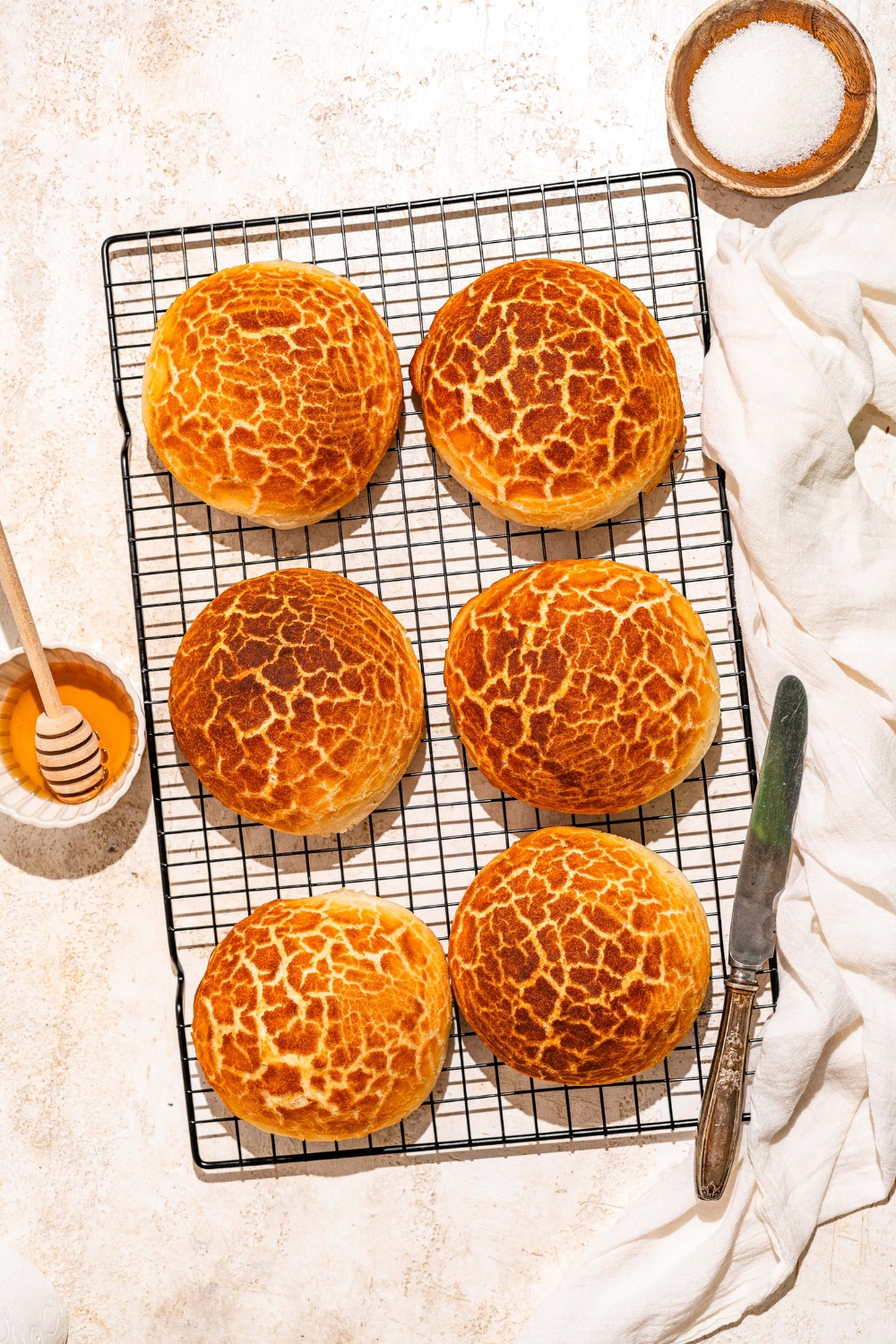 A wire rack with baked Dutch Crunch bread loaves. The rack is on a tan counter with a knife and bowl of honey.