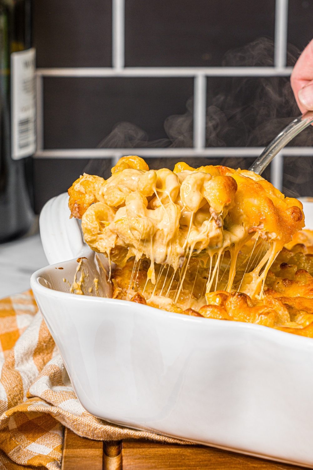 A baking pan with baked bacon macaroni and cheese with a spoon taking a scoop from the pan. The pan is on a wooden board with a yellow checkered napkin.