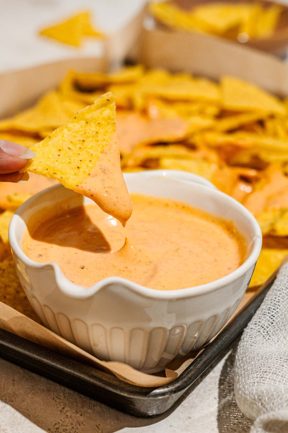 A baking sheet lined with parchment paper with nacho chips and a bowl of cheddar cheese sauce. A chip is being dipped into the sauce.