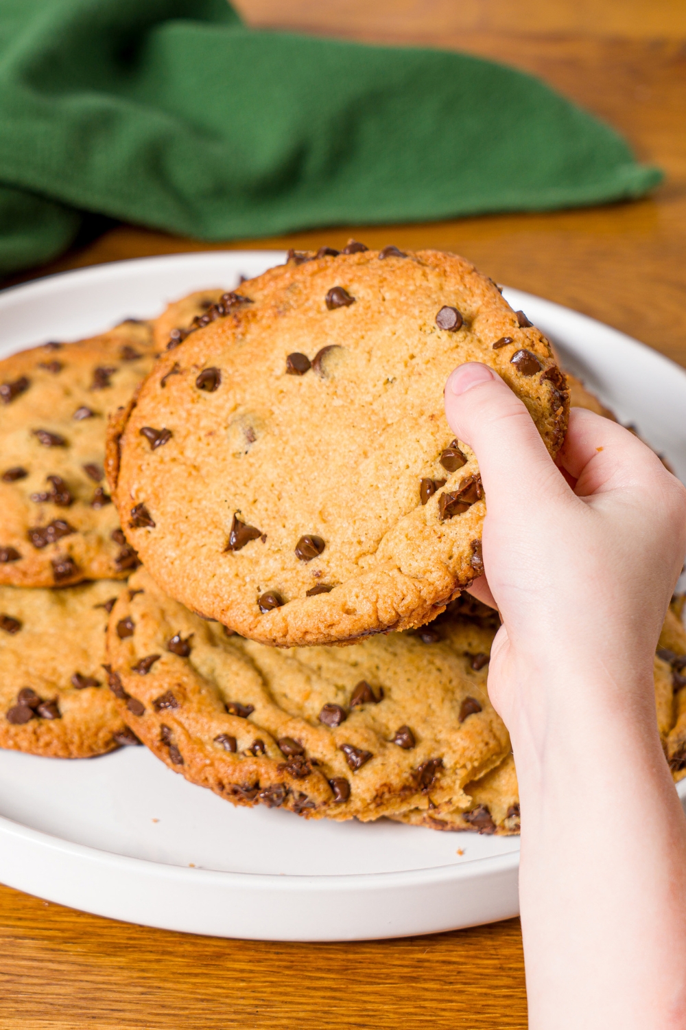 A white plate with chocolate chip cookies without baking soda. A hand is taking a cookie from the plate. The plate is on a wooden counter with a green cloth napkin.