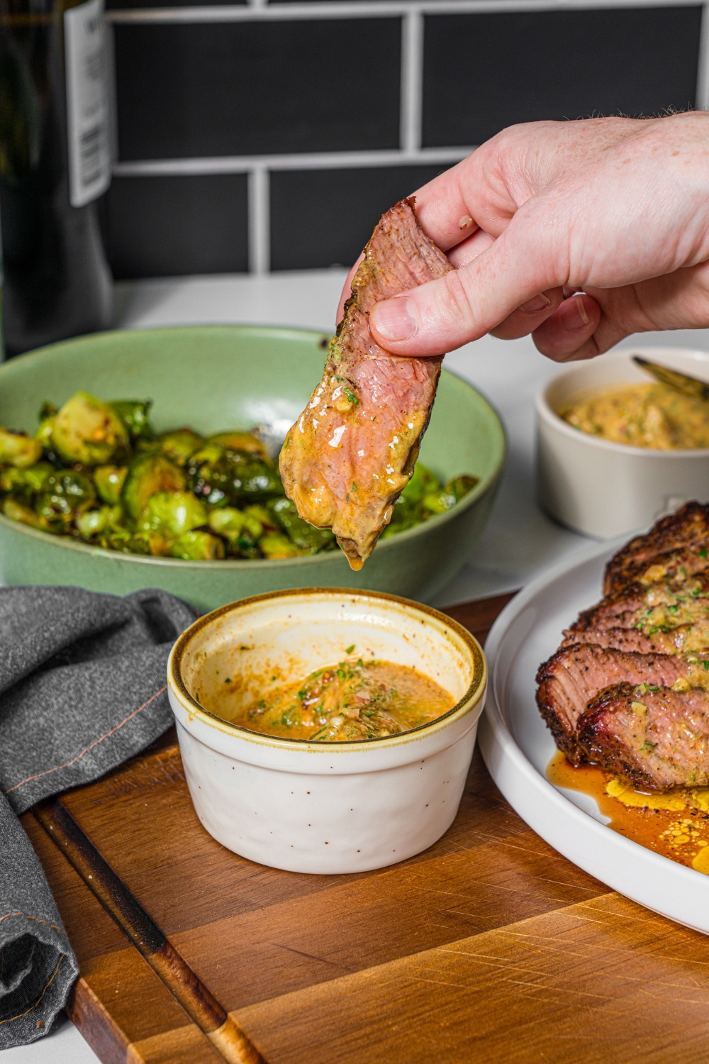 A hand dipping a slice of steak into a bowl of cowboy butter. The bowl is on a wooden board with a plate of steak and a bowl of roasted brussels sprouts.