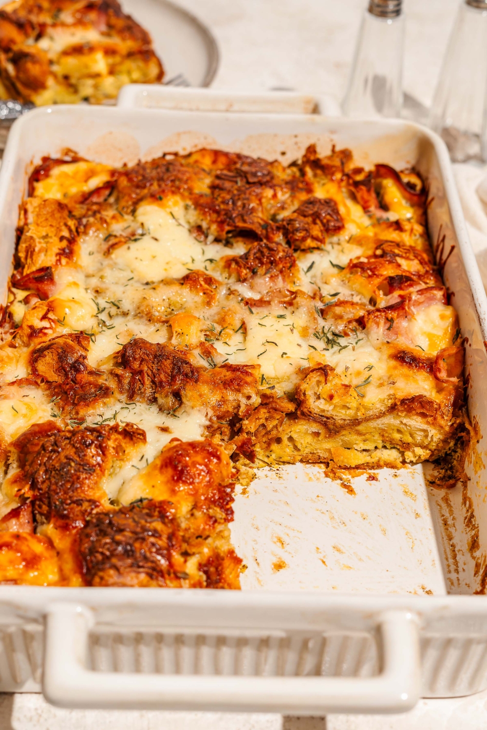 A baking dish with croissant breakfast casserole with ham and cheese. A slice is taken from the casserole. The dish is on a tan counter with a plate of casserole.