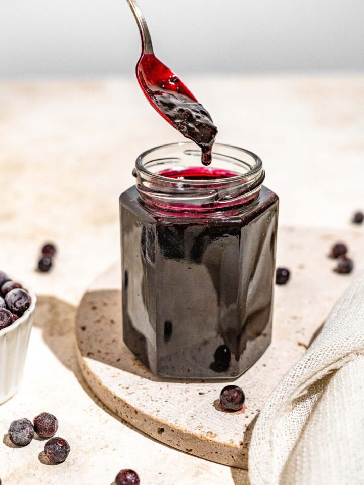 A jar of elderberry jam with a spoon dipping in the jar. The jar is on a marble plate on a tan counter with a bowl of elderberries.