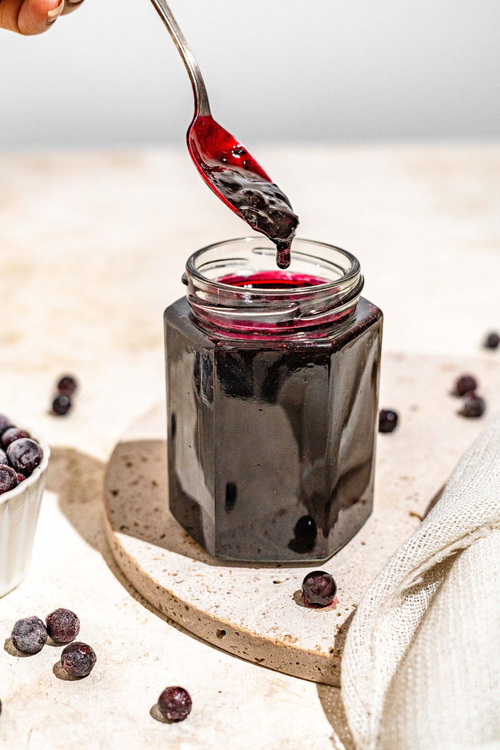 A jar of elderberry jam with a spoon dipping in the jar. The jar is on a marble plate on a tan counter with a bowl of elderberries.