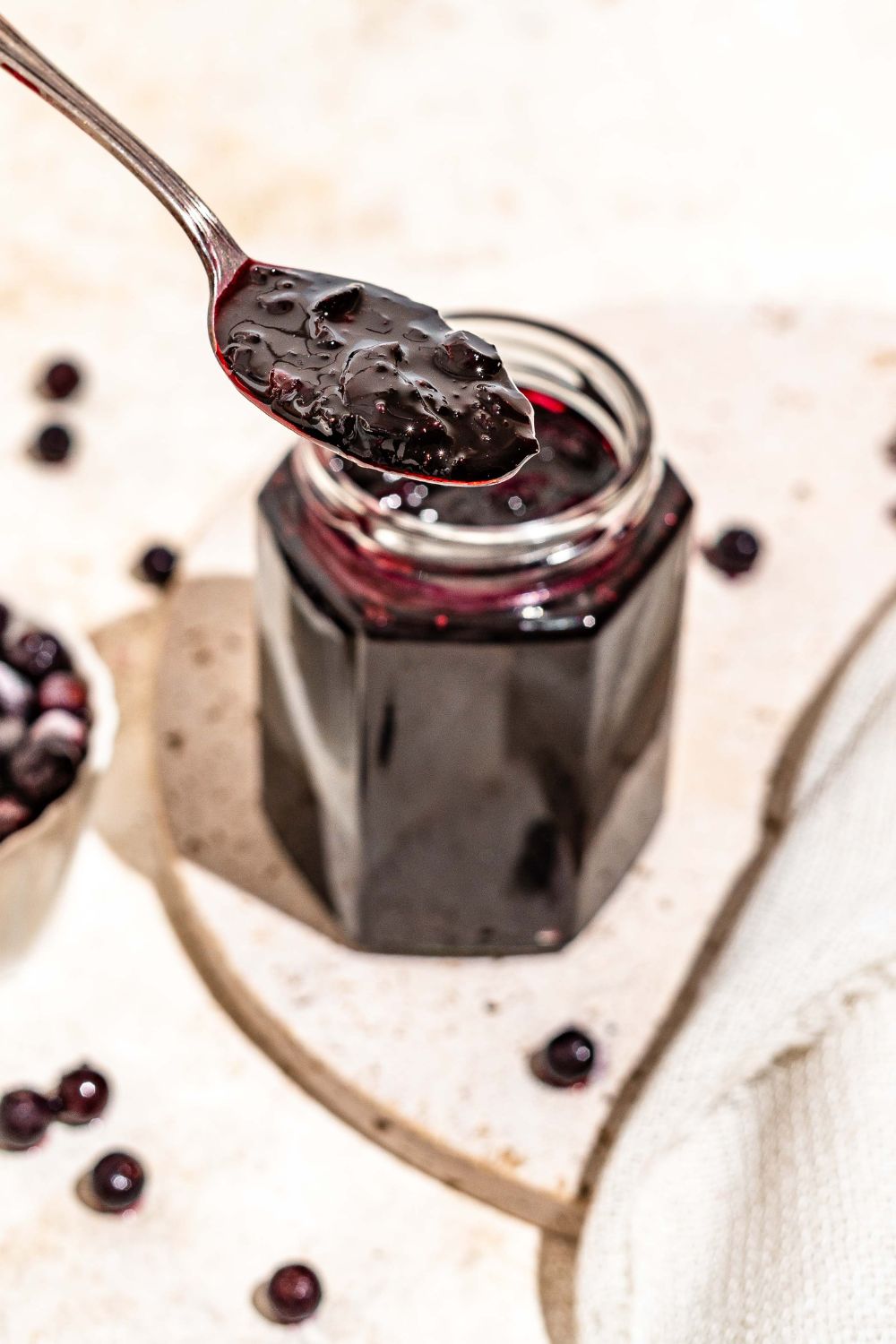 A jar of elderberry jam with a spoon dipping in the jar. The jar is on a marble plate on a tan counter with a bowl of elderberries.