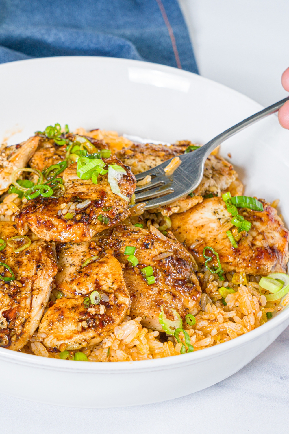 A white bowl with sliced seasoned chicken breast served over rice with sliced green onion. A fork is taking a bite from the bowl.