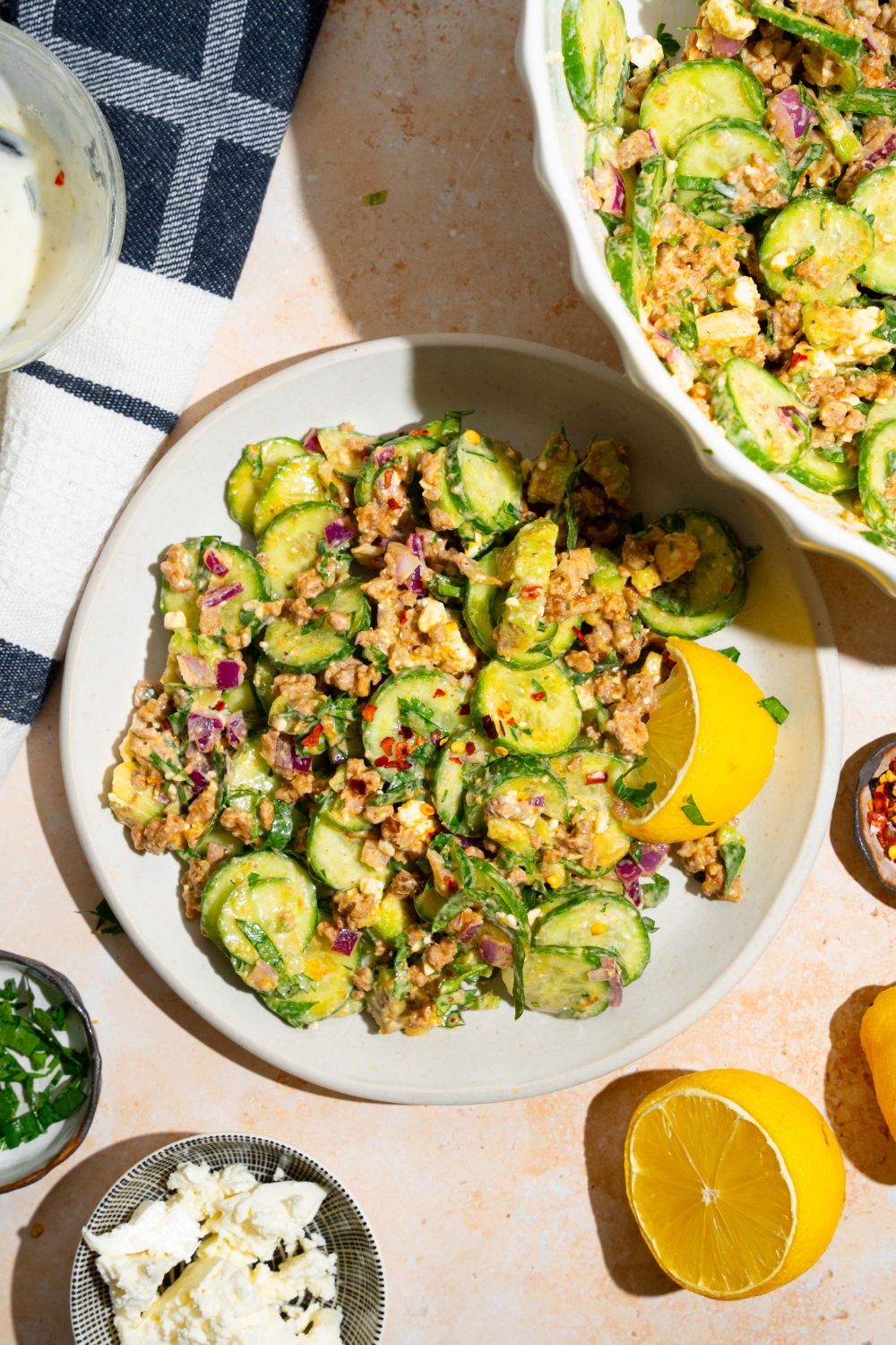 A white plate with high protein cucumber salad with cucumber, red onion, avocado, seasoned ground beef, and feta cheese tossed in greek yogurt dressing. The salad is served with a lemon wedge. The plate is on a tan counter with small bowls of ingredients and a large bowl of cucumber salad.