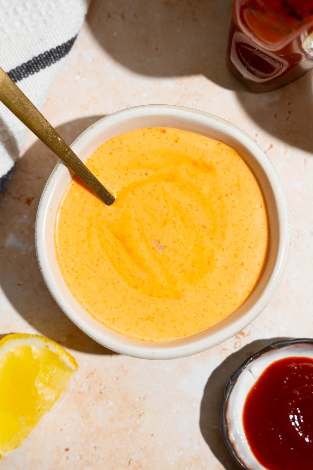 A bowl of honey sriracha sauce with a spoon in the bowl. The bowl is on a tan counter with a jar of honey and bowl of sriracha.