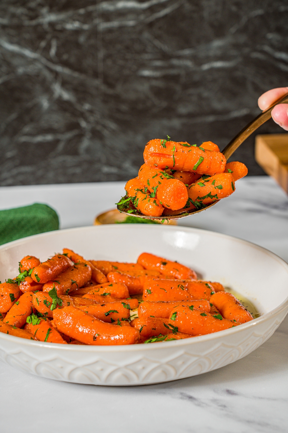 A bowl of honey roasted baby carrots garnished with fresh parsley with a spoon with a bite of carrots. The bowl is on a marble counter.