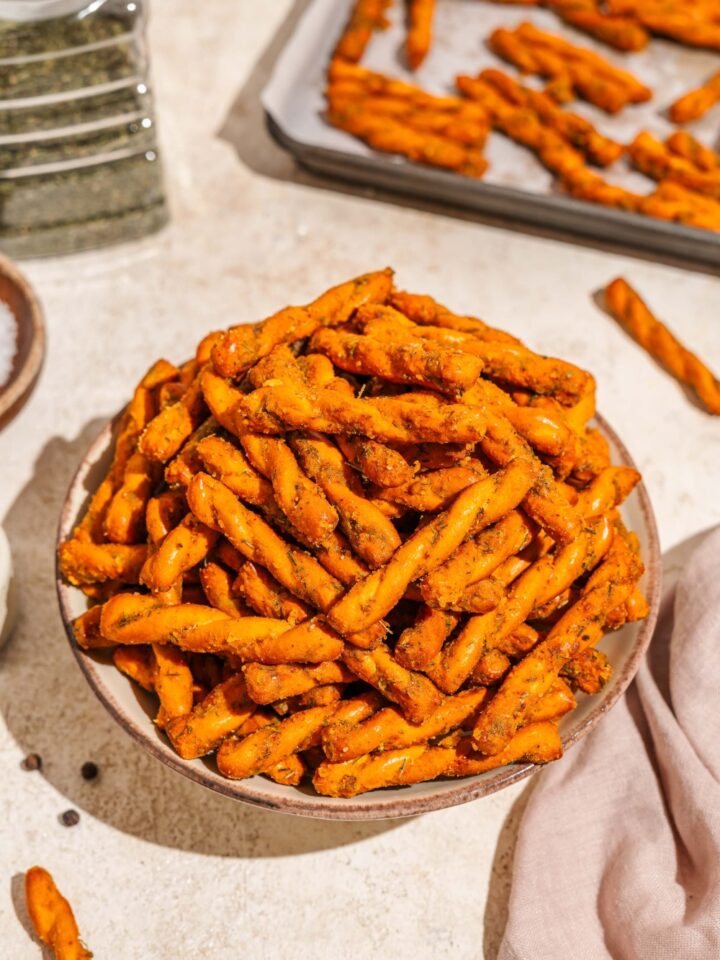 A bowl of seasoned pretzel braids. The bowl is on a tan counter with a white cloth napkin and a baking sheet with pretzels.
