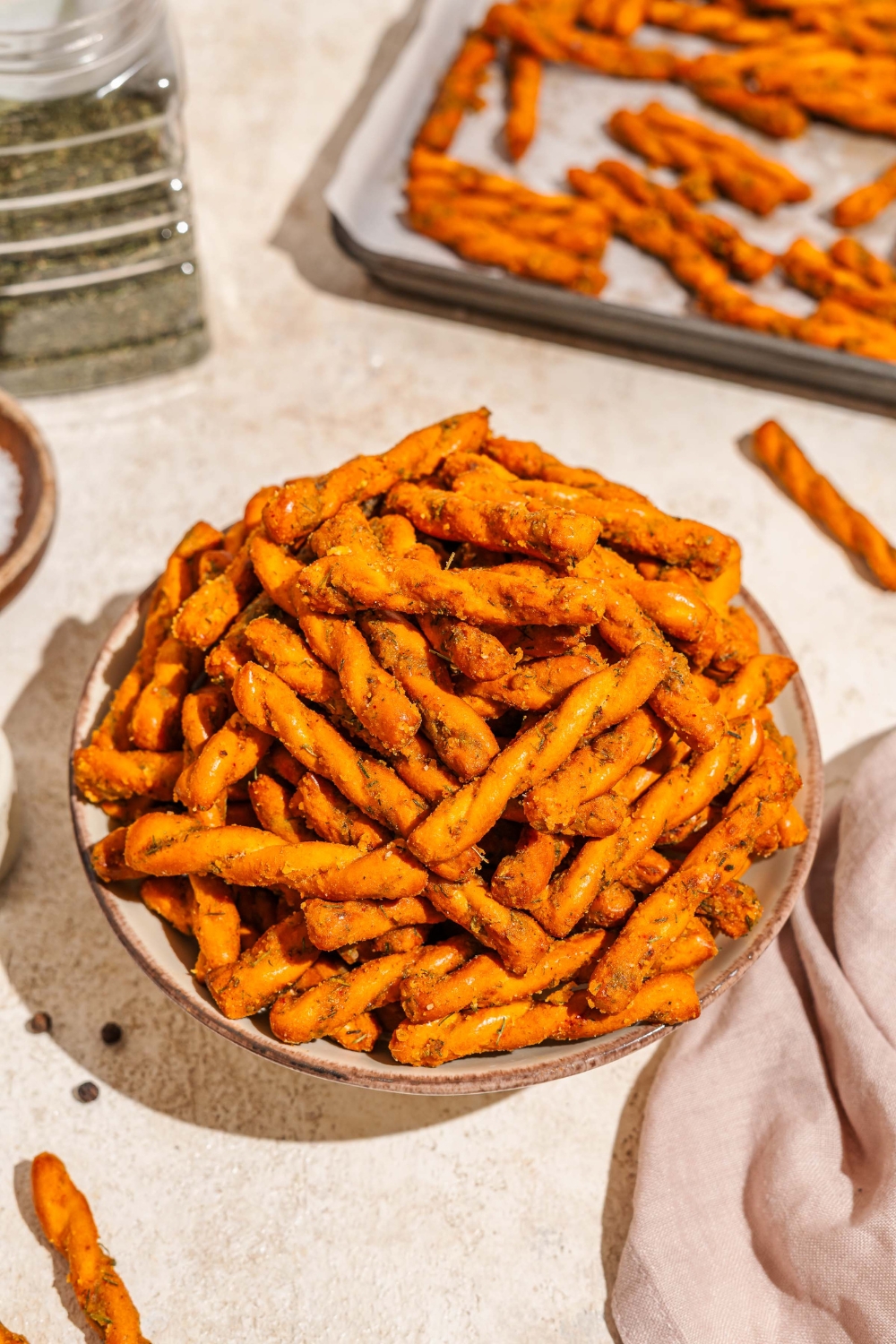 A bowl of seasoned pretzel braids. The bowl is on a tan counter with a white cloth napkin and a baking sheet with pretzels.