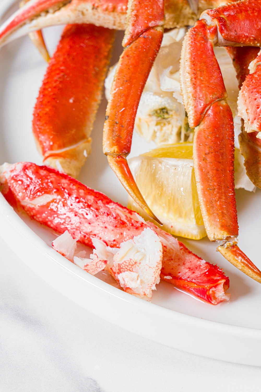 A white plate with a piece of crab leg meat with steamed snow crab legs served with lemon.