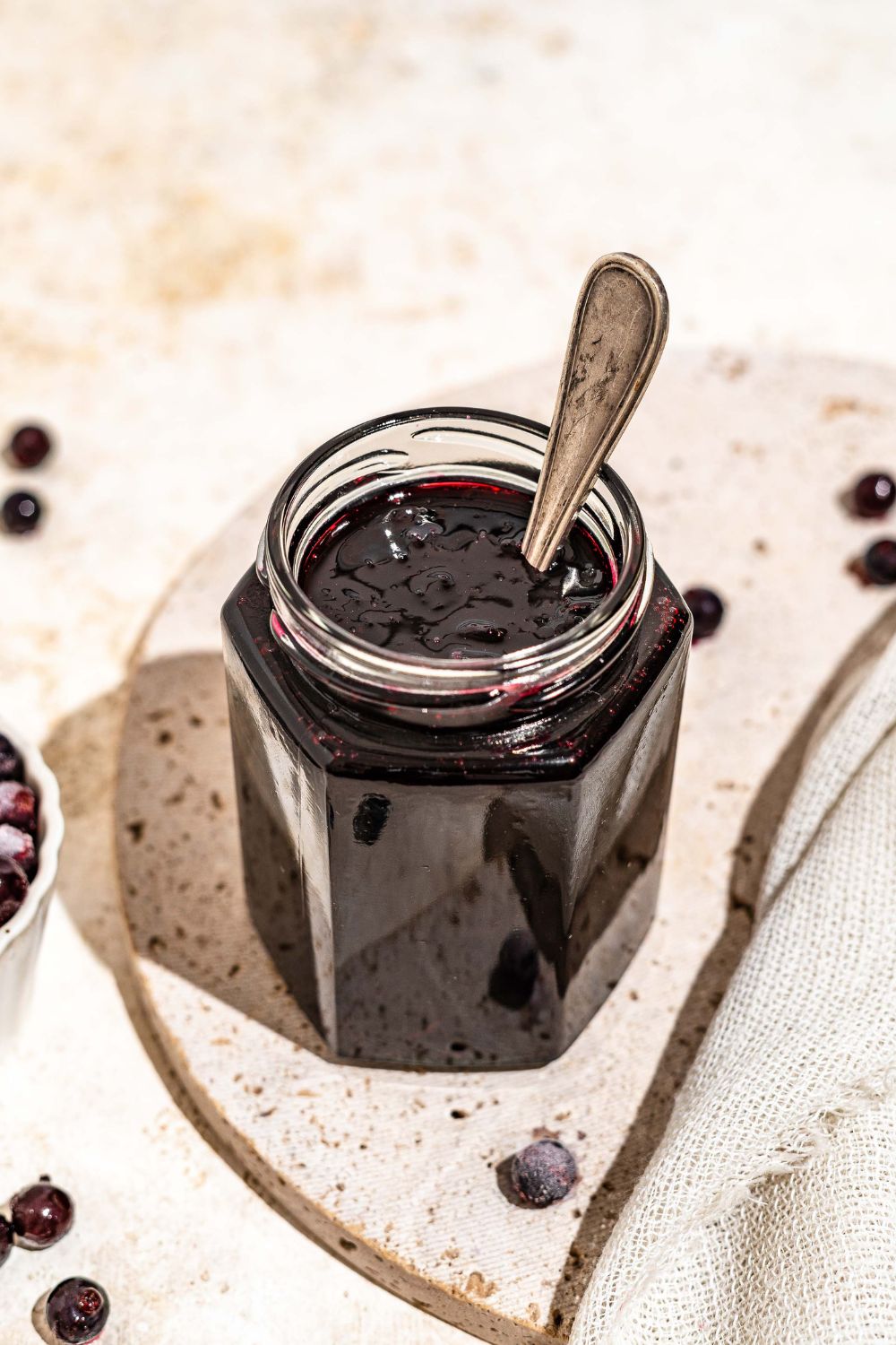 A jar of elderberry jam with a spoon in the jar. The jar is on a marble plate on a tan counter with a bowl of elderberries.