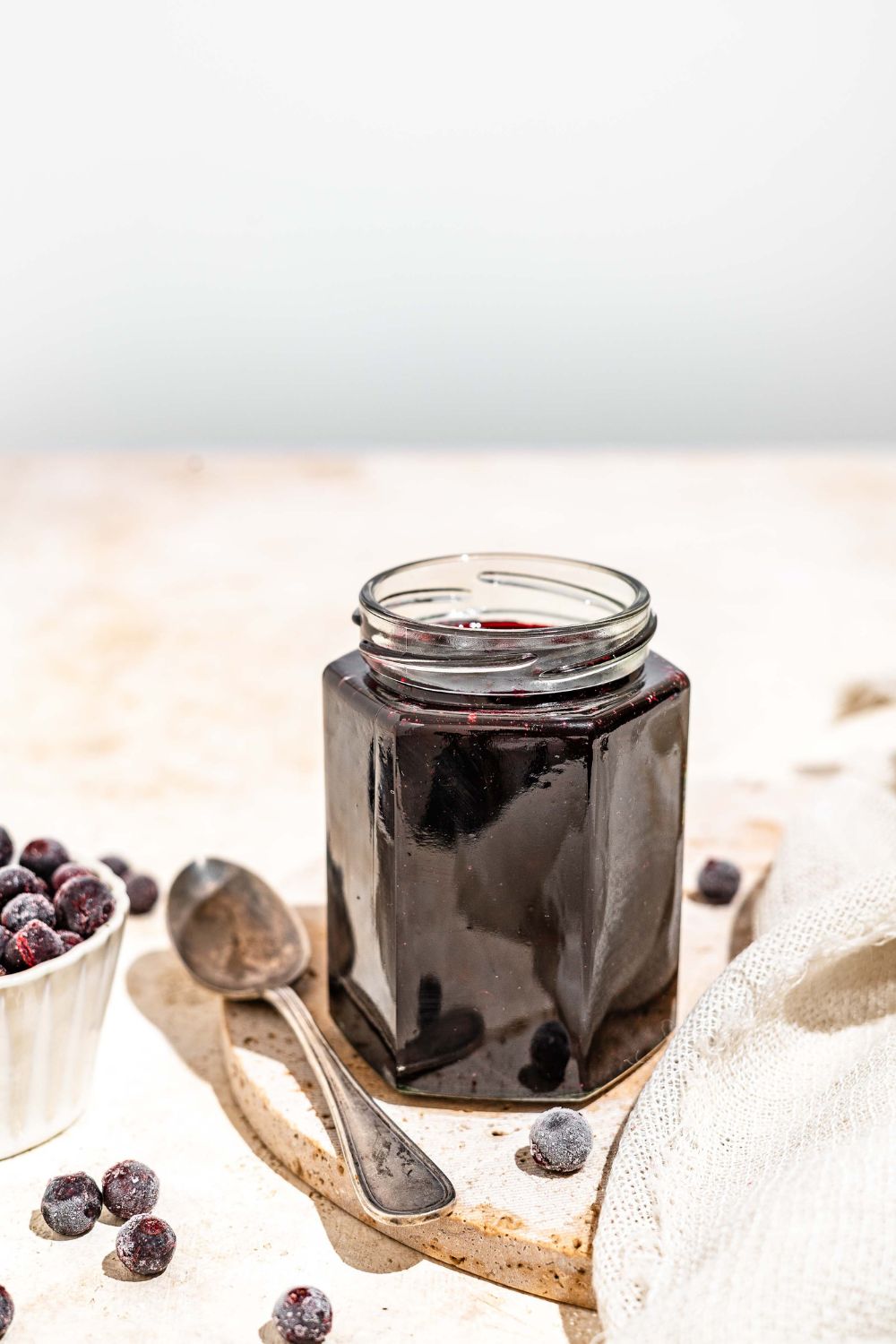 A jar of elderberry jam with a spoon next to it. The jar is on a marble plate on a tan counter with a bowl of elderberries.
