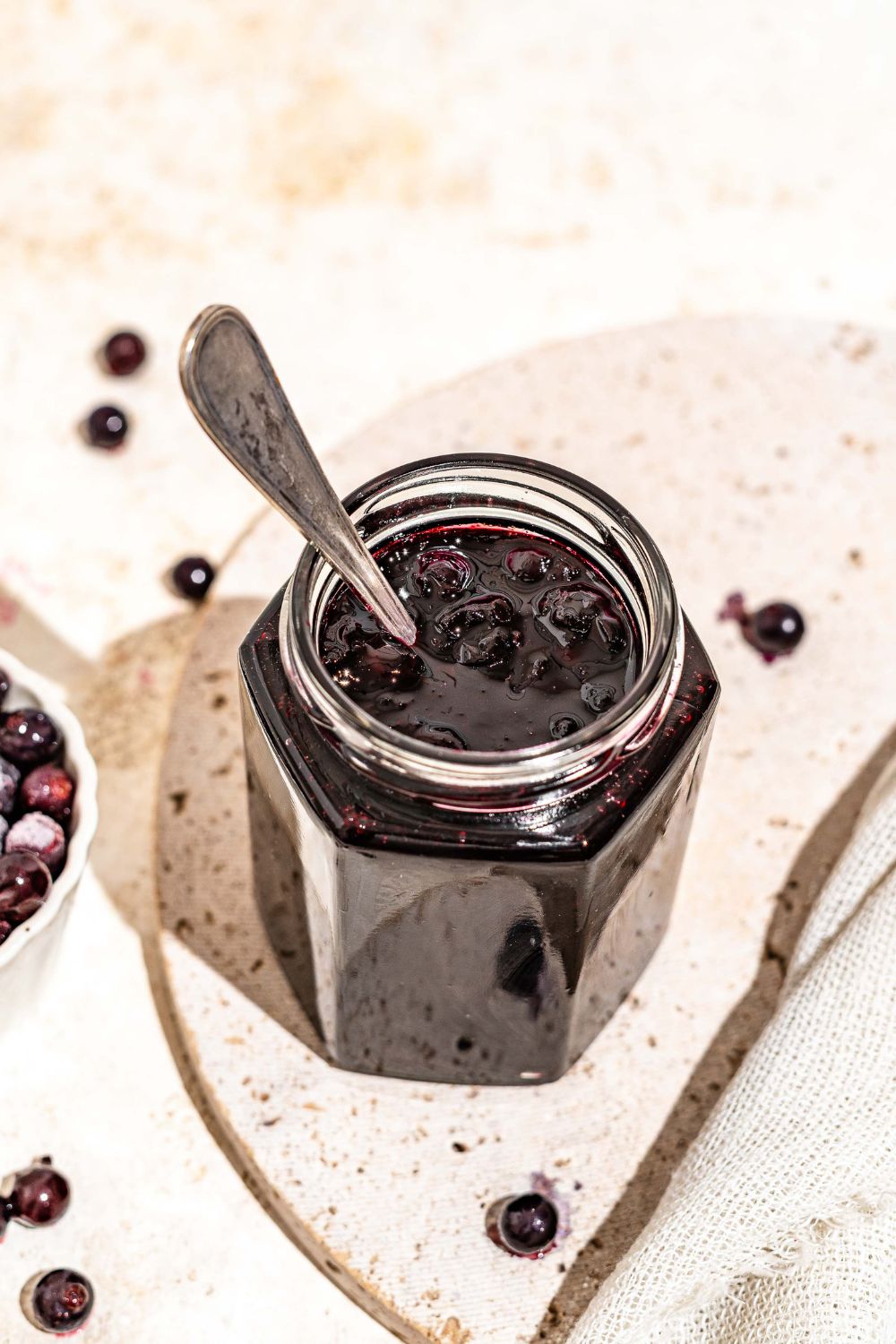 A jar of elderberry jam with a spoon in the jar. The jar is on a marble plate on a tan counter with a bowl of elderberries.