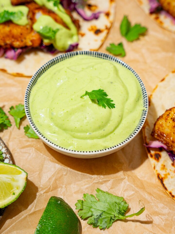 A bowl of fish taco sauce on a tan counter lined with parchment paper with several open fish tacos.