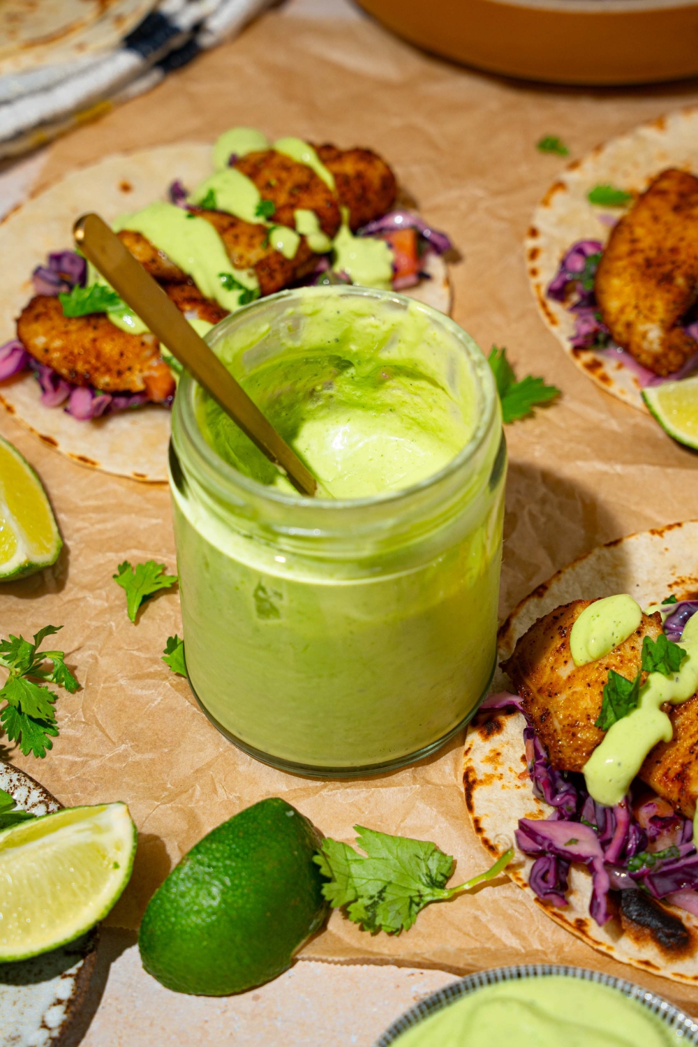 A jar of fish taco sauce on a tan counter lined with parchment paper with several open fish tacos. There is a spoon in the jar.