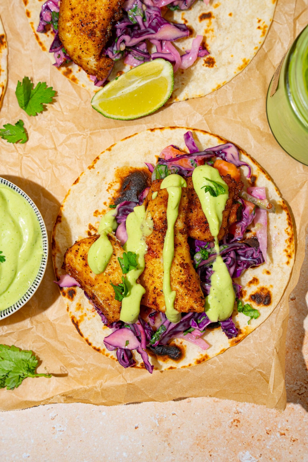 A tan counter with parchment paper with several open fish tacos topped with cabbage and drizzled with fish taco sauce. There is a bowl of taco sauce on the counter.