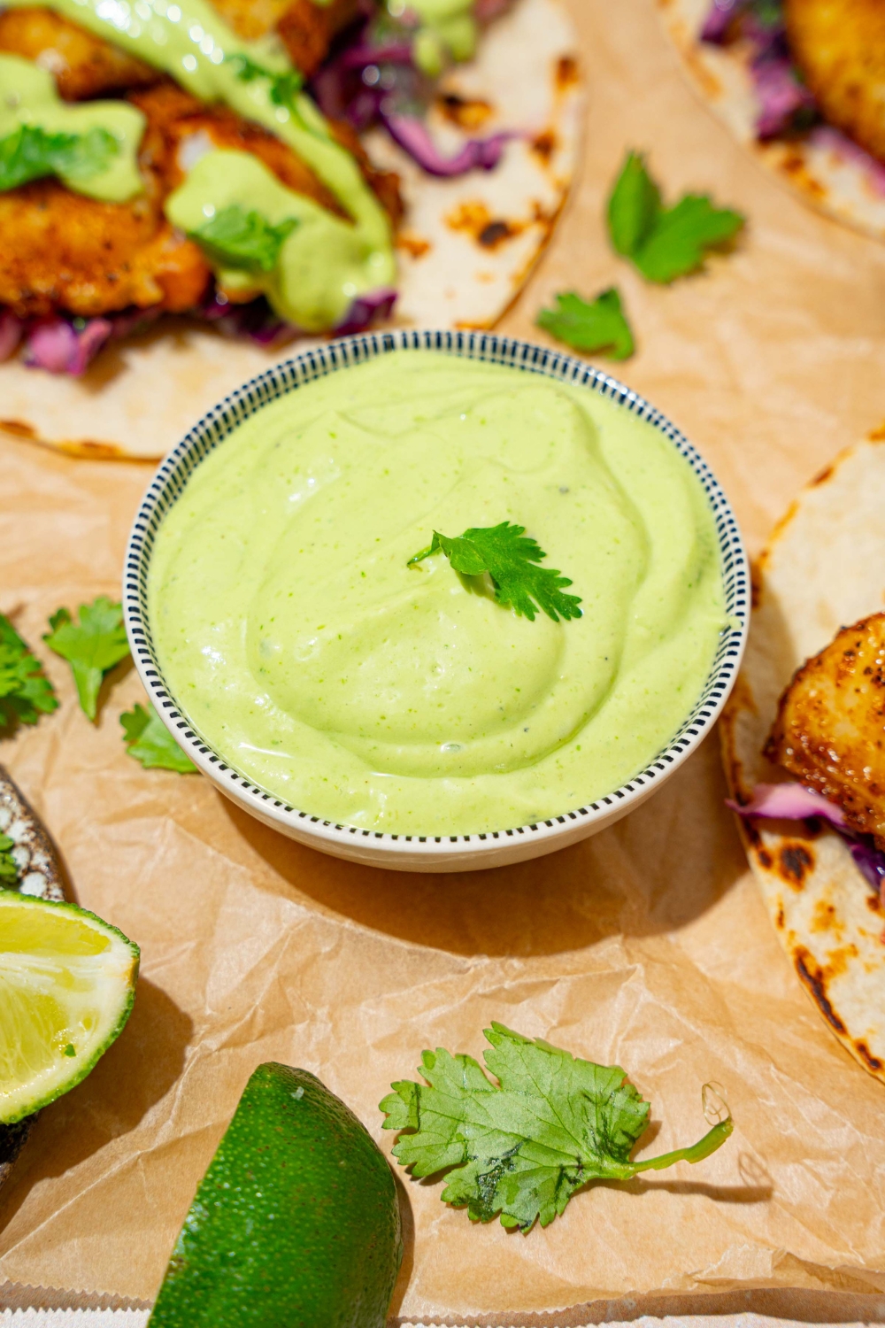 A bowl of fish taco sauce on a tan counter lined with parchment paper with several open fish tacos.