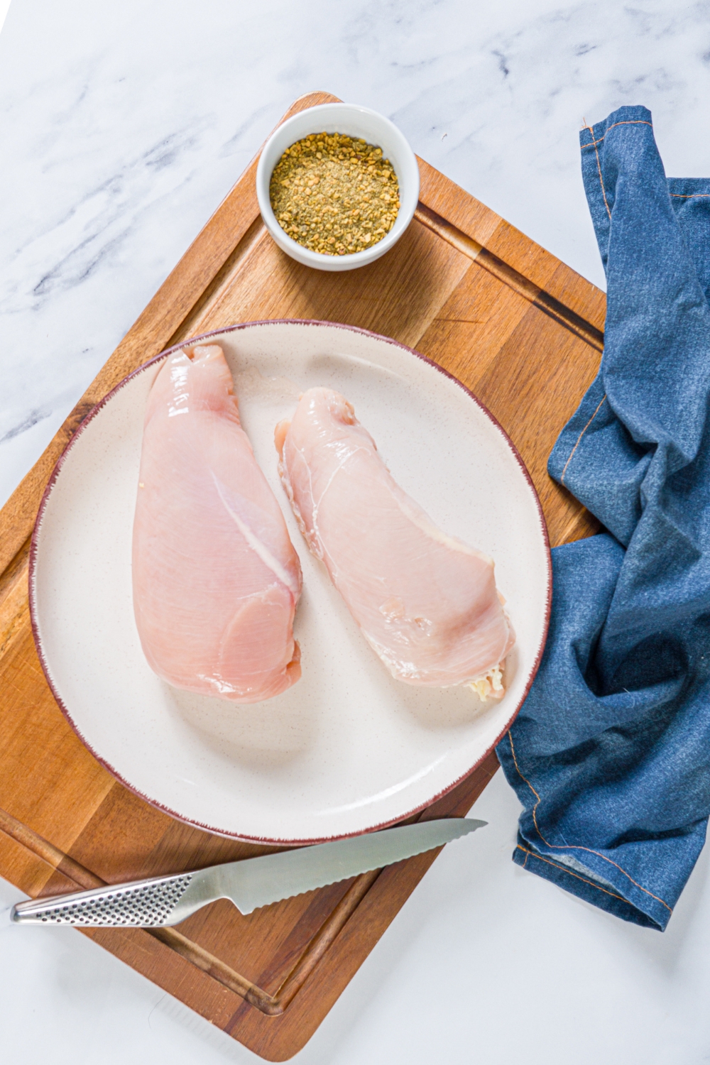 A wooden cutting board with a plate of defrosted chicken breasts and a bowl of seasonings. The board is on a marble counter with a blue cloth napkin.