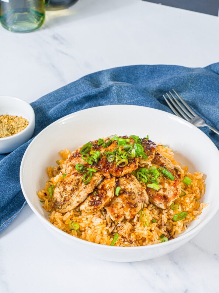 A white bowl with sliced seasoned chicken breast served over rice with sliced green onion. The bowl is on a marble counter with a blue cloth napkin and fork.
