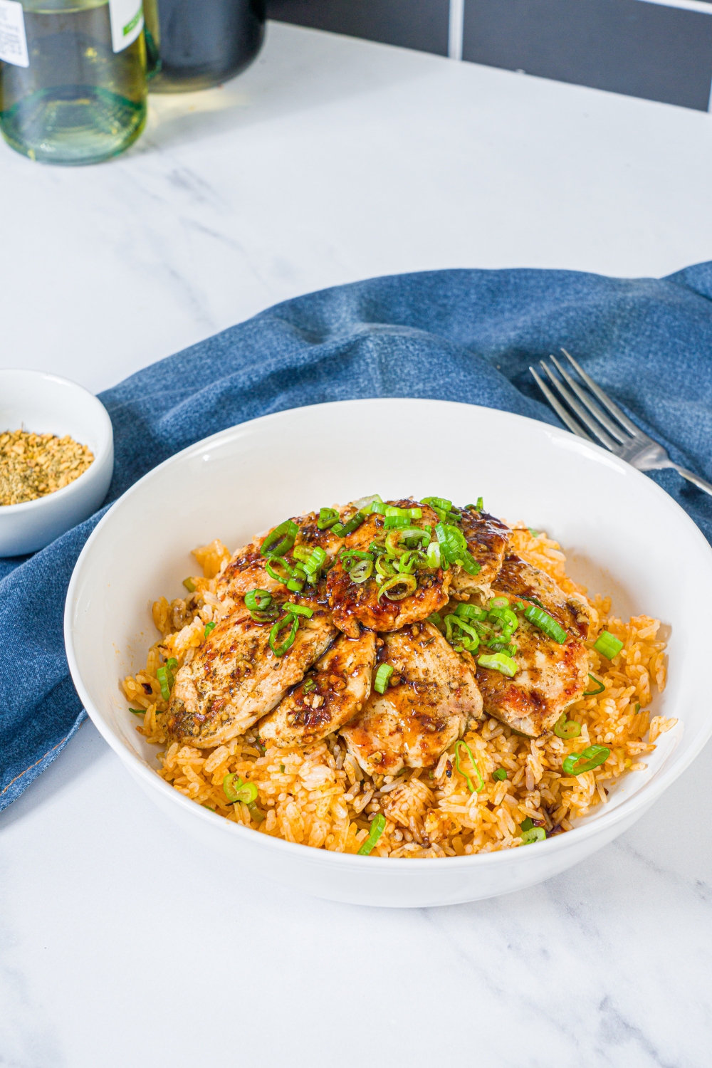A white bowl with sliced seasoned chicken breast served over rice with sliced green onion. The bowl is on a marble counter with a blue cloth napkin and fork.
