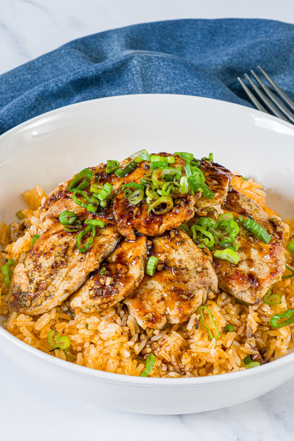A white bowl with sliced seasoned chicken breast served over rice with sliced green onion. The bowl is on a marble counter with a blue cloth napkin and fork.