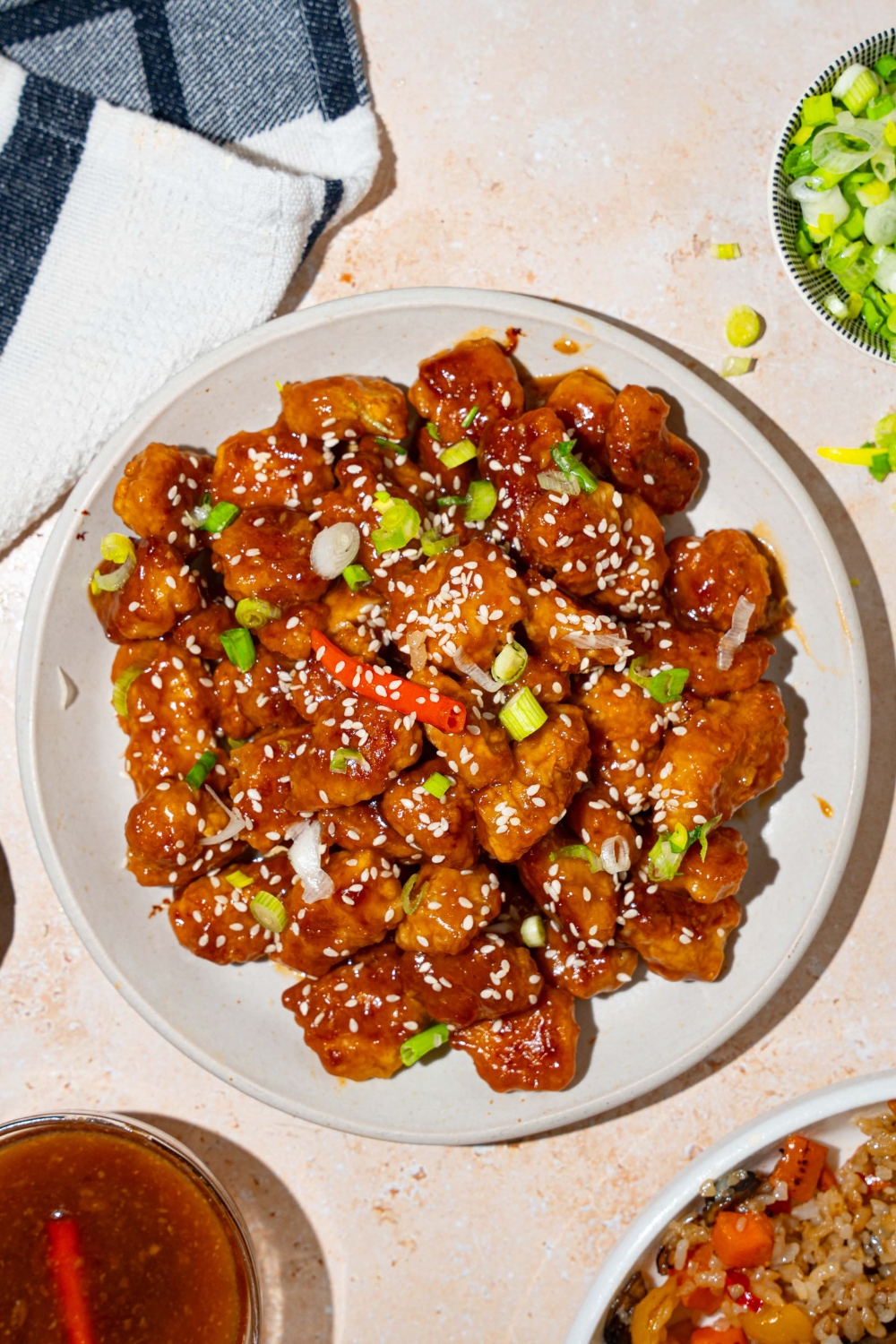 A white plate with general tso's chicken garnished with sesame seeds, sliced green onion, and red chile. The plate is on a tan counter with a bowl of sauce.