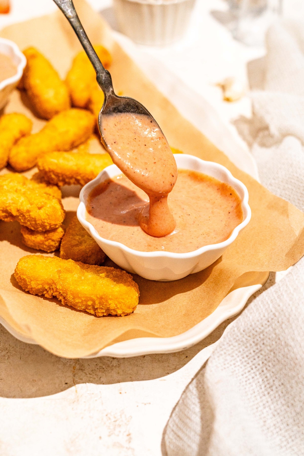 A white platter lined with parchment paper with fried fish sticks and a bowl of gochujang mayo. A spoon is dipping into the bowl. The platter is on a tan counter.