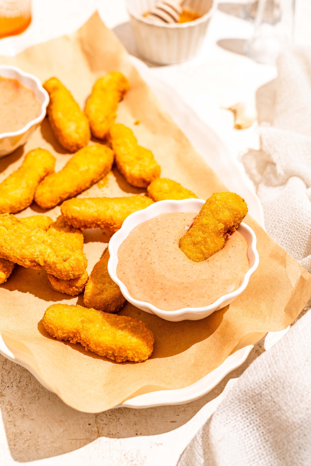 A white platter lined with parchment paper with fried fish sticks and bowls of gochujang mayo. A fish stick is dipped in the bowl. The platter is on a tan counter.
