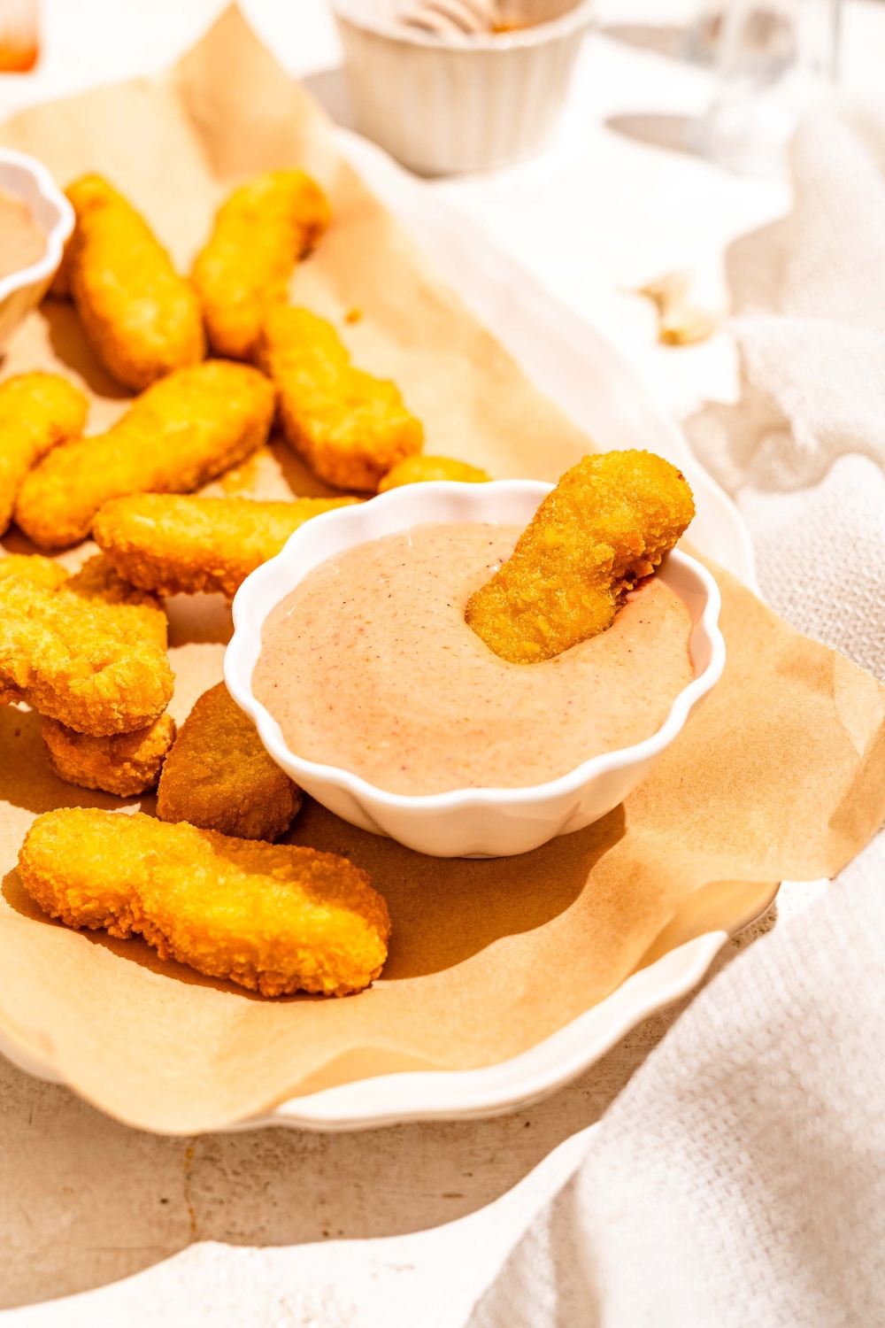 A white platter lined with parchment paper with fried fish sticks and bowls of gochujang mayo. A fish stick is dipped in the bowl. The platter is on a tan counter.