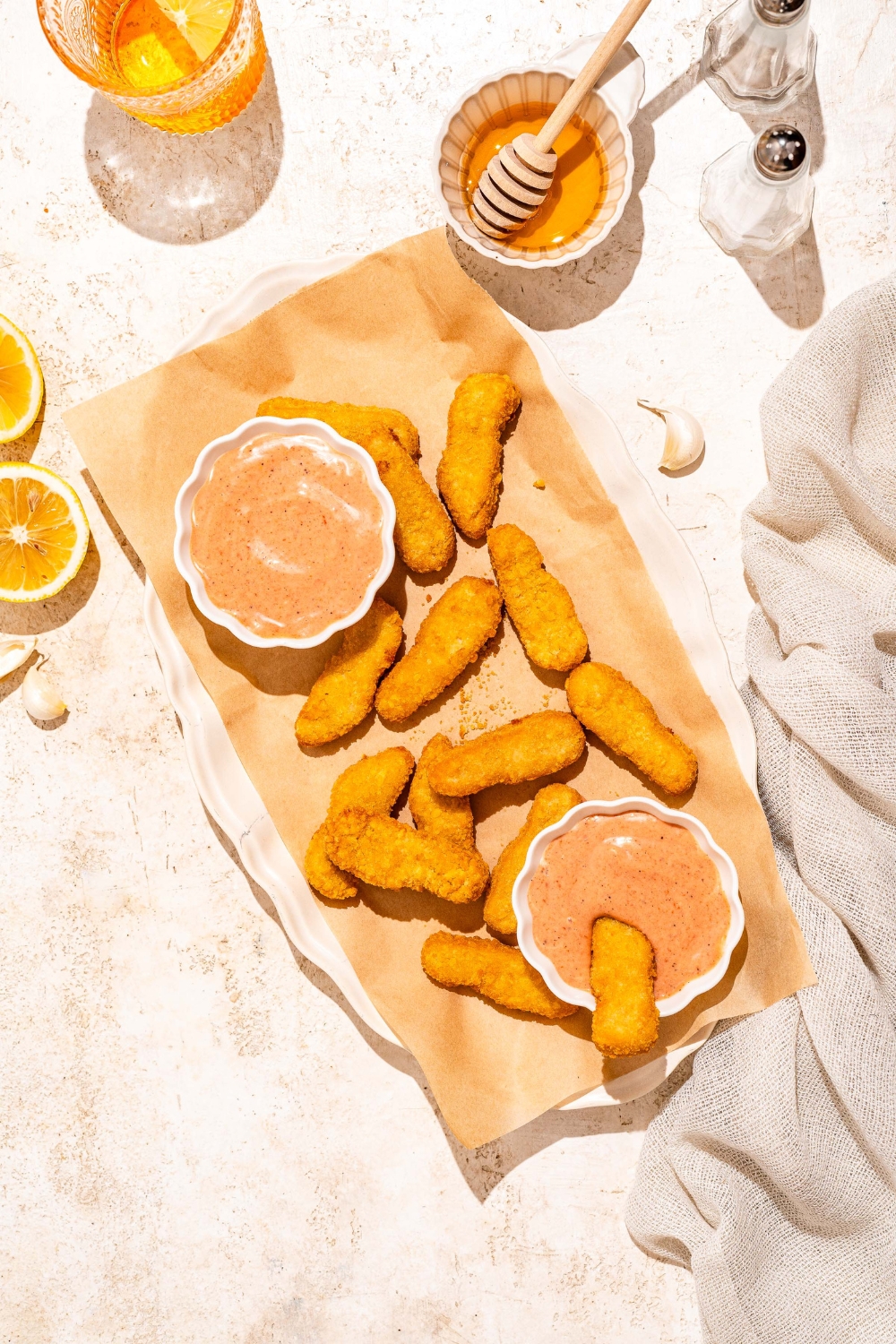 A white platter lined with parchment paper with fried fish sticks and bowls of gochujang mayo. A fish stick is dipped in one of the bowls. The platter is on a tan counter with a cloth napkin and bowl of honey.