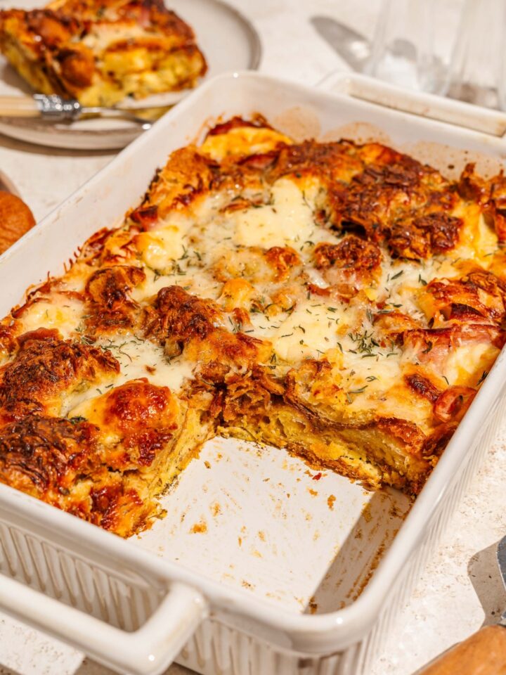 A baking dish with croissant breakfast casserole with ham and cheese. A slice is taken from the casserole. The dish is on a tan counter with a plate of casserole.