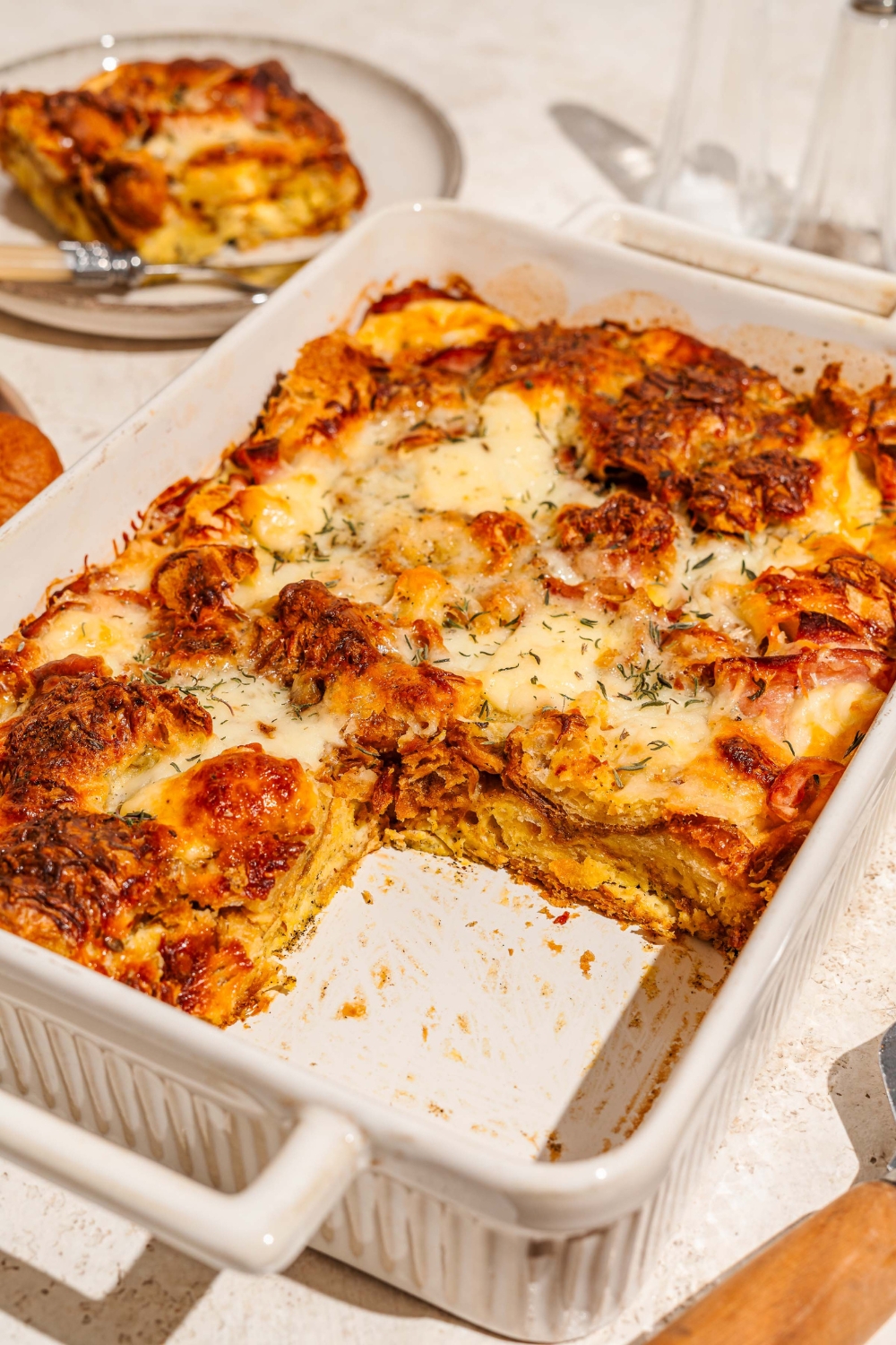 A baking dish with croissant breakfast casserole with ham and cheese. A slice is taken from the casserole. The dish is on a tan counter with a plate of casserole.