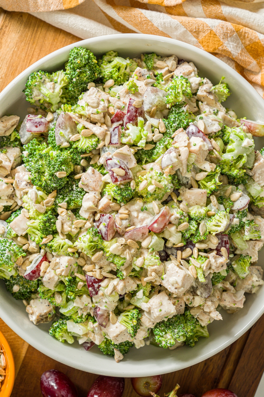 A large bowl of broccoli chicken salad with sunflower seeds and grapes, tossed in a greek yogurt dressing. The bowl is on a wooden board with a yellow checkered napkin.