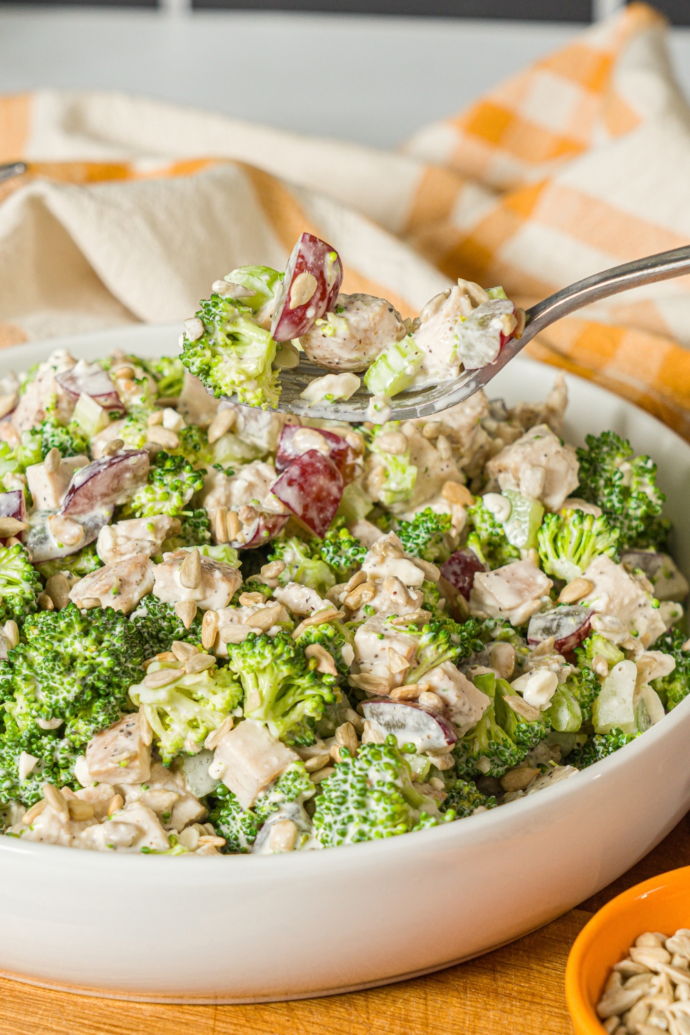 A large bowl of broccoli chicken salad with sunflower seeds and grapes, tossed in a greek yogurt dressing. A fork is taking a bite from the bowl. The bowl is on a wooden board with a yellow checkered napkin.