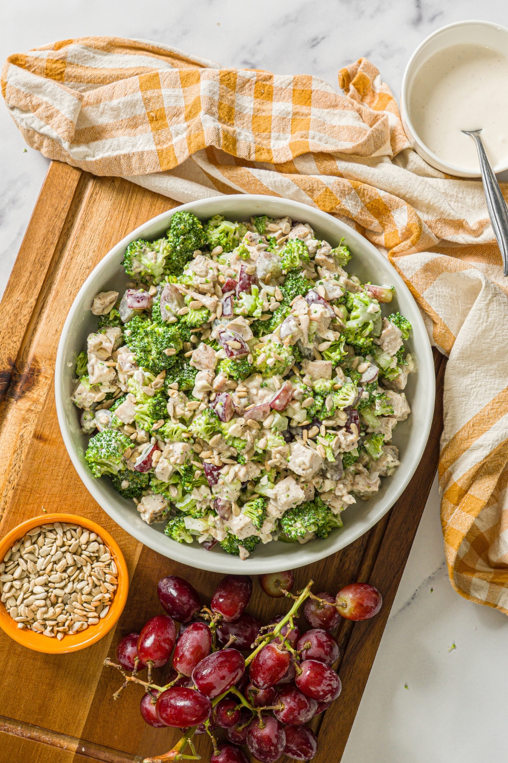 A large bowl of broccoli chicken salad with sunflower seeds and grapes, tossed in a greek yogurt dressing. The bowl is on a wooden board with a yellow checkered napkin and bowl of sunflower seeds.