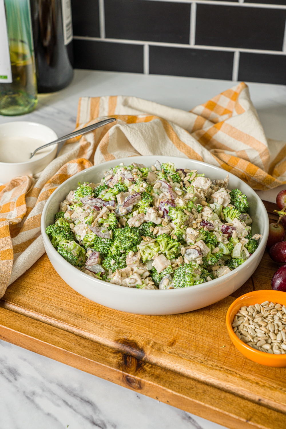 A large bowl of broccoli chicken salad with sunflower seeds and grapes, tossed in a greek yogurt dressing. The bowl is on a wooden board with a yellow checkered napkin and bowl of sunflower seeds.