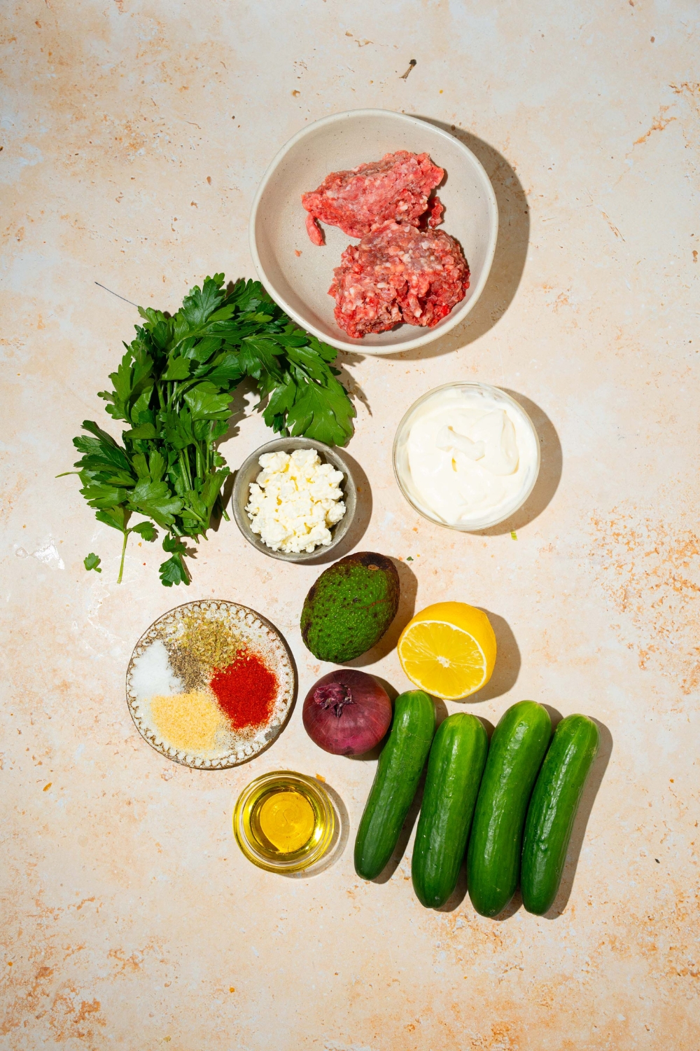 A tan counter with ingredients to make high protein cucumber salad including ground beef, cucumber, feta cheese, avocado, lemon, red onion, oil, greek yogurt, and seasonings.