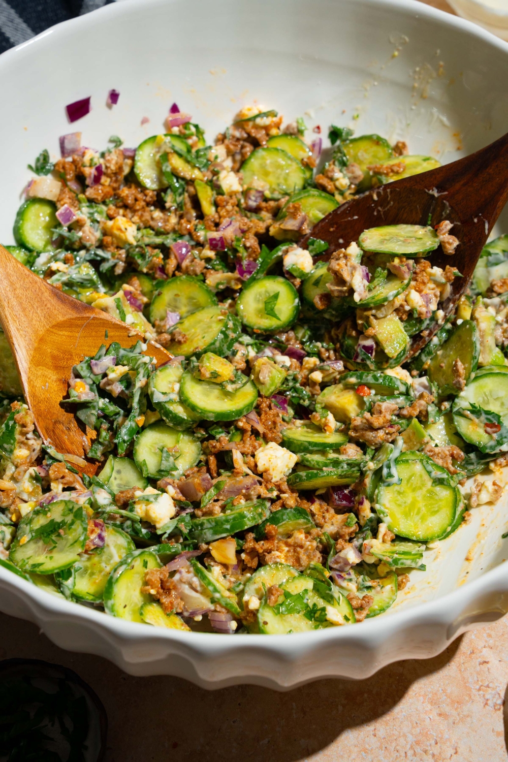 A large white bowl with two salad tongs tossing a high protein cucumber salad including cucumbers, feta cheese, avocado, red onion, and greek yogurt dressing. The bowl is on a tan counter.