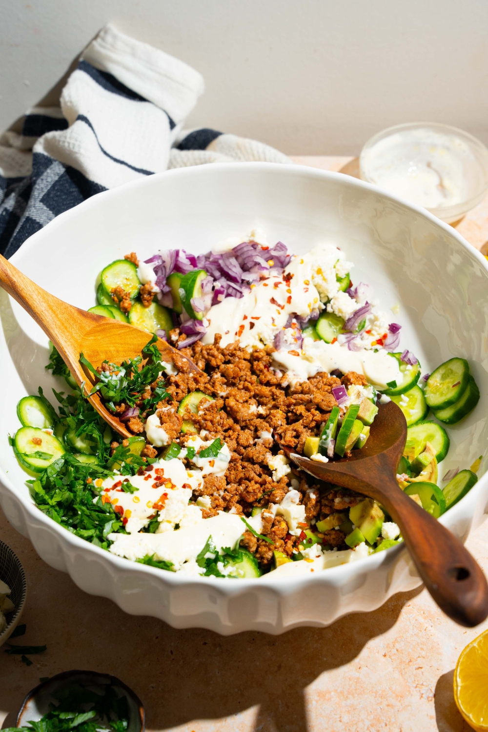 A large bowl with high protein cucumber salad ingredients piled with two salad tongs. The bowl contains cucumbers, feta cheese, avocado, seasoned ground beef, red onion, and greek yogurt dressing. The bowl is on a tan counter.