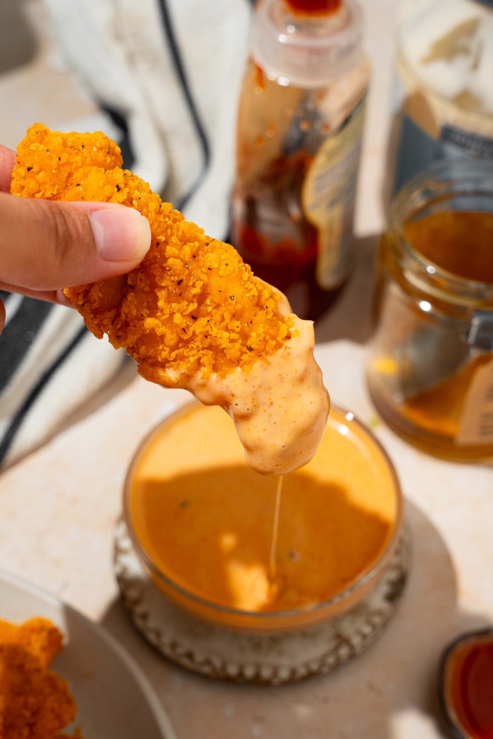A close up of a chicken tender dipped in bang bang sauce. There is a bowl of sauce on a saucer on a tan counter blurred in the background.