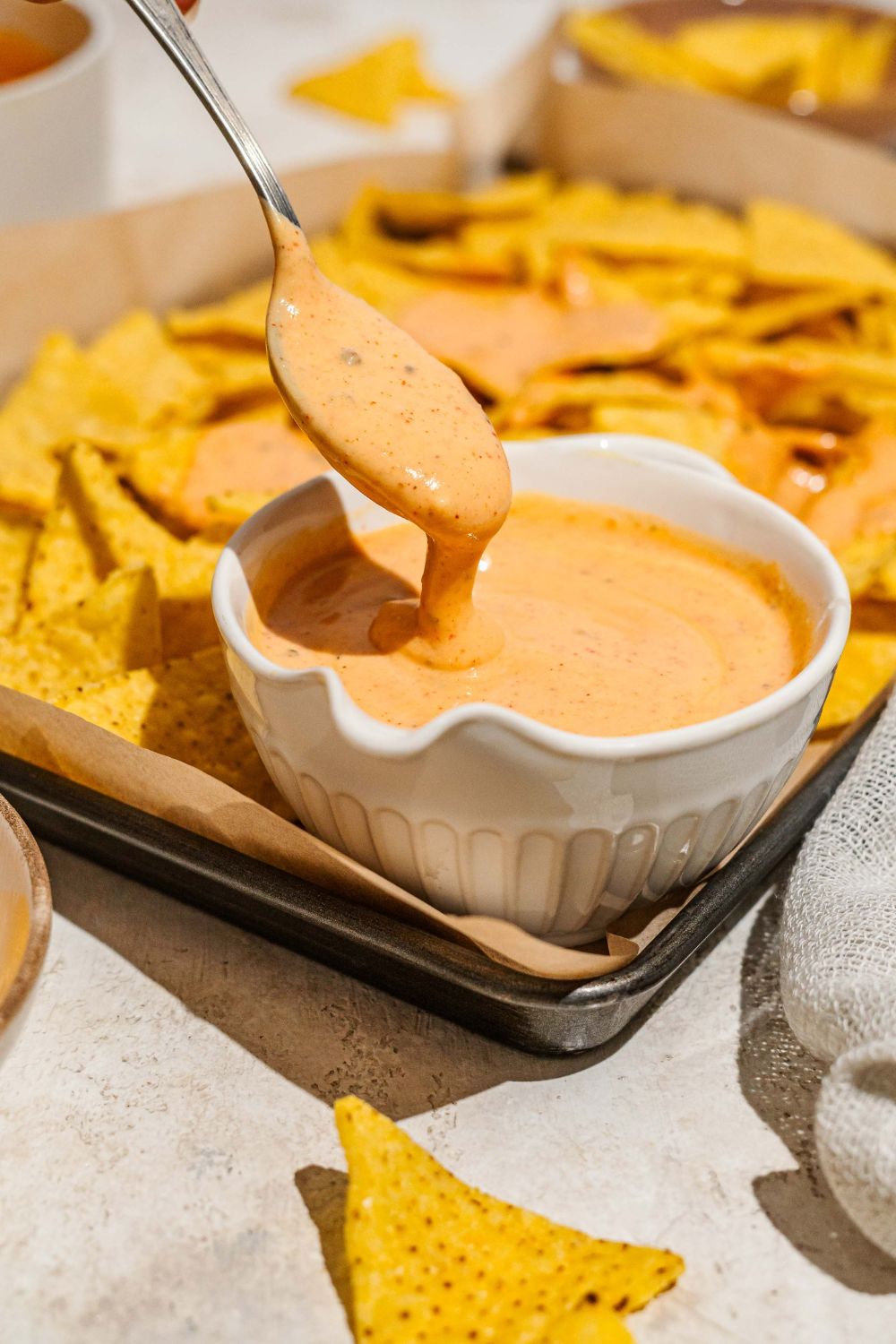 A baking sheet lined with parchment paper with nacho chips and a bowl of cheddar cheese sauce. A spoon is dipping in the cheese.