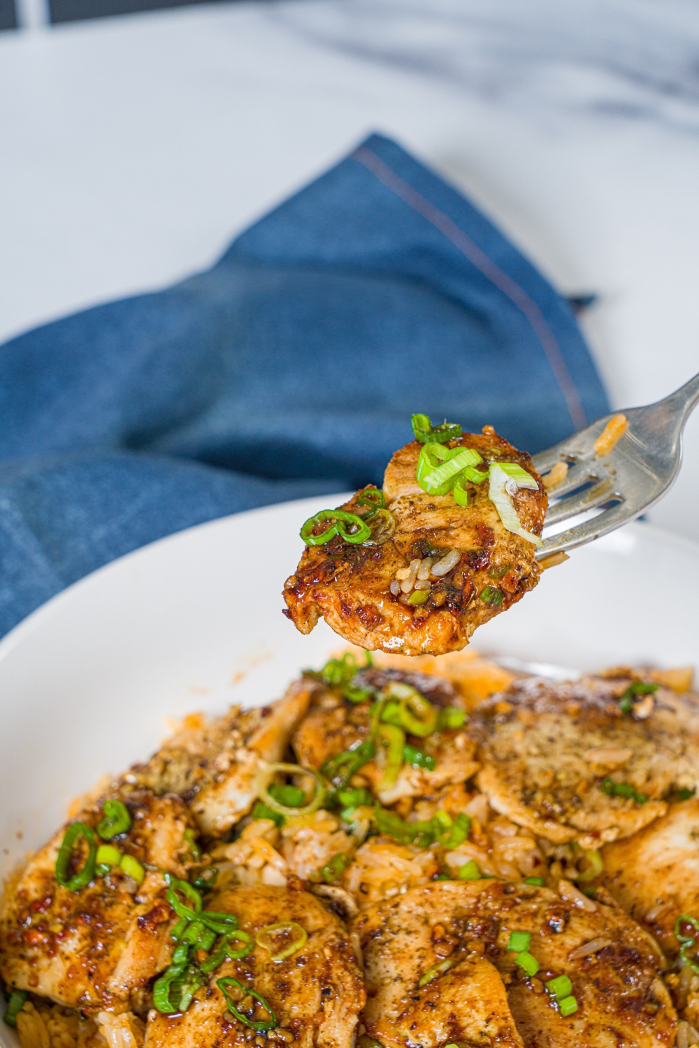 A white bowl with sliced seasoned chicken breast served over rice with sliced green onion. A fork is taking a bite from the bowl.