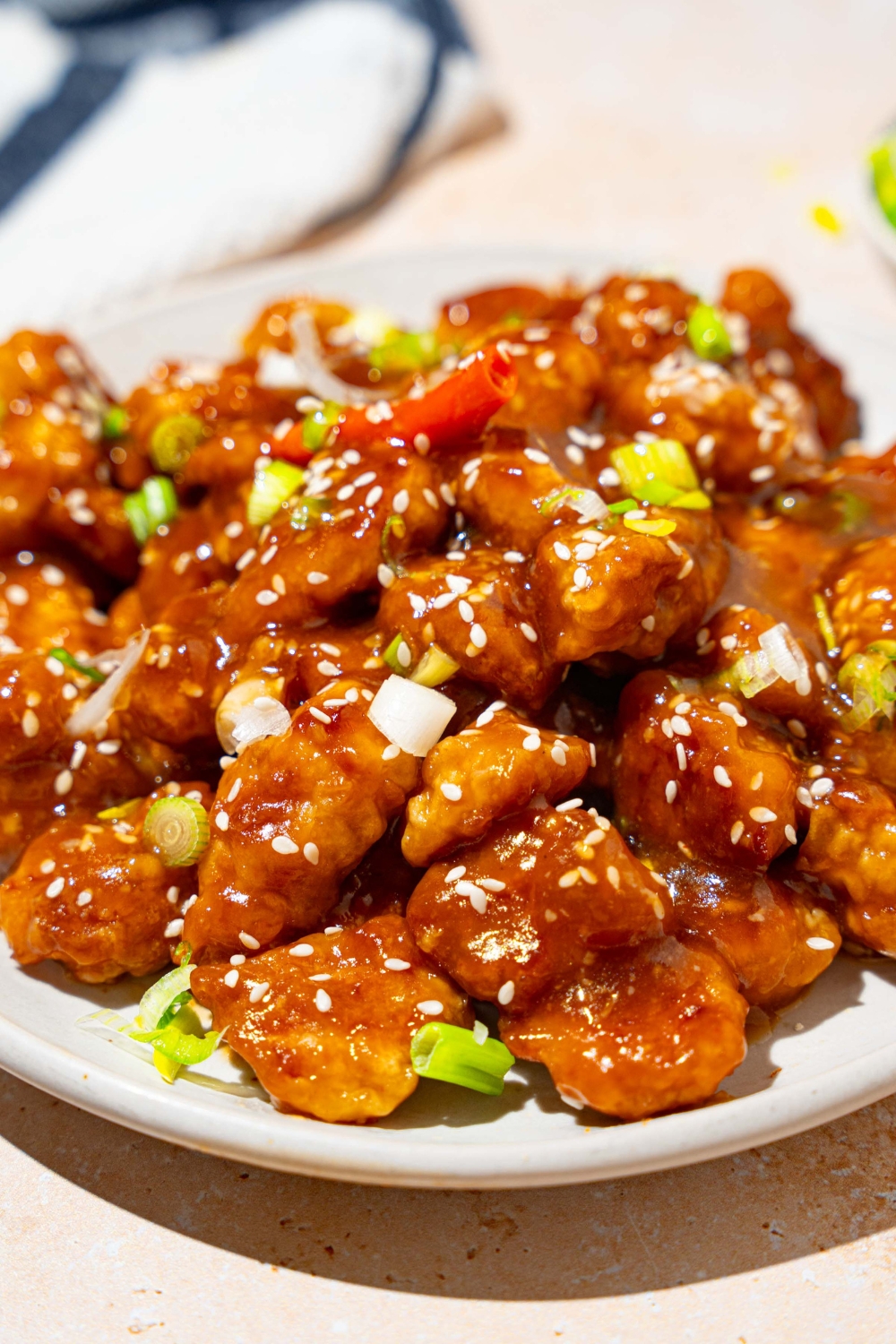 A white plate with general tso's chicken garnished with sesame seeds, sliced green onion, and red chile. The plate is on a tan counter.